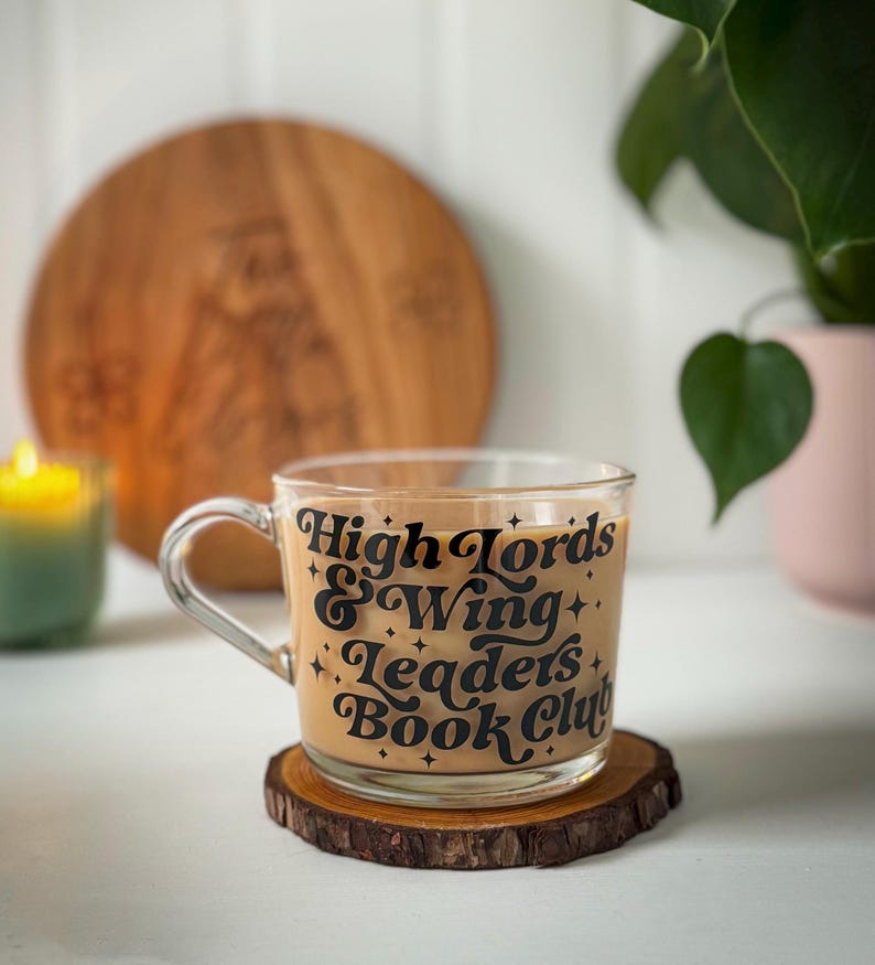 May include: A clear glass mug filled with a light brown drink, inscribed with "High Lords & Wing Leaders Book Club" in black lettering. The mug rests on a small, round wooden coaster. A lit green candle and a wooden board are in the background.