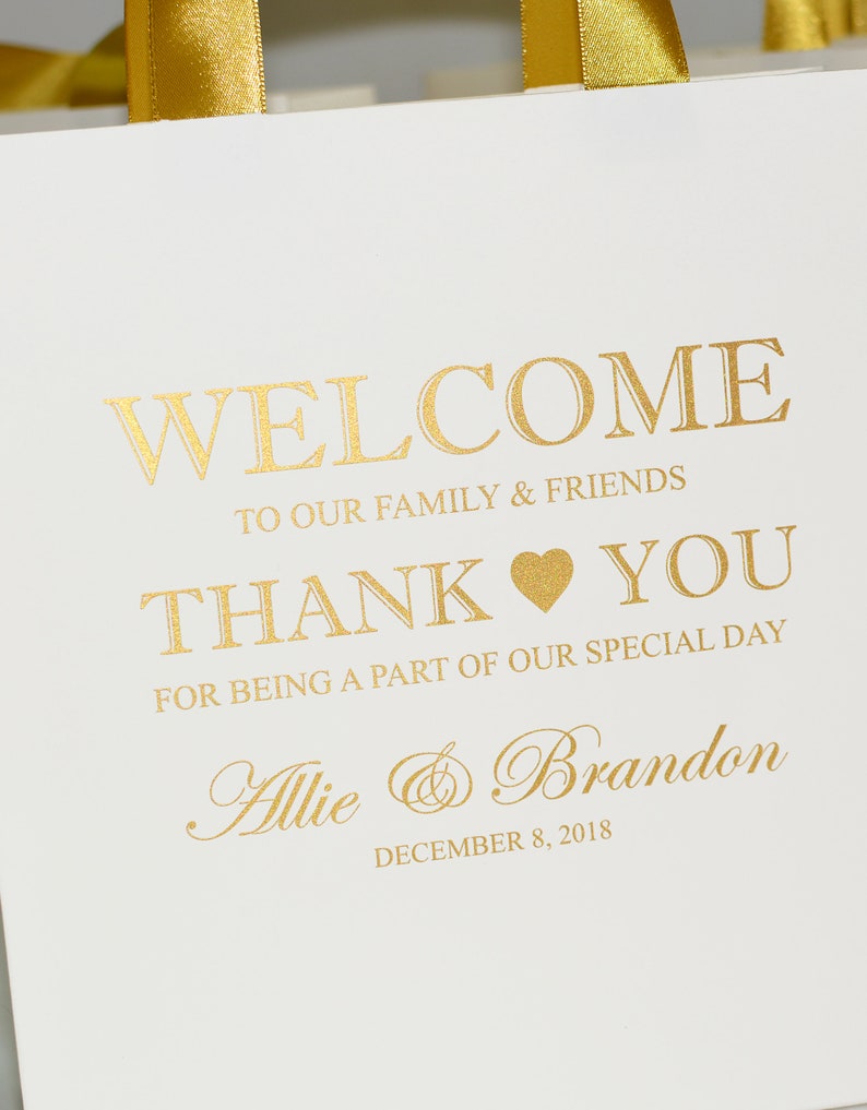 May include: White favor bag with gold ribbon handles. The bag has gold lettering that says "Welcome to our family & friends Thank You for being a part of our special day Allie & Brandon December 8, 2018".