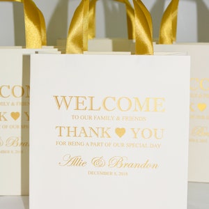 May include: White favor bags with gold ribbon handles and gold lettering. The bags say "Welcome to our family & friends, Thank you for being a part of our special day, Allie & Brandon, December 8, 2018".