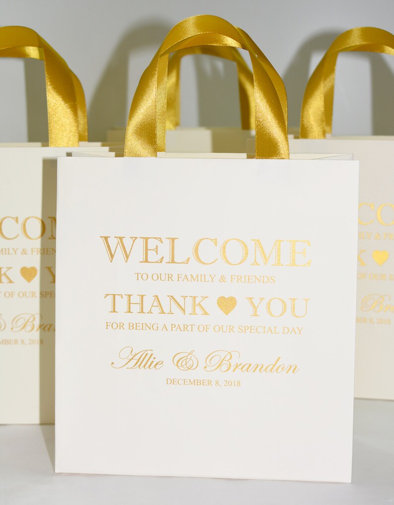 May include: White favor bags with gold ribbon handles and gold lettering. The bags say "Welcome to our family & friends, Thank you for being a part of our special day, Allie & Brandon, December 8, 2018".