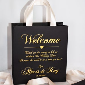 May include: Black gift bag with white satin handles. The bag has gold lettering that says "Welcome", "Thank you for coming to help us celebrate Our Wedding Day! It means the world to us to have you here! Love, Alexis & Roy 10.24.2020".