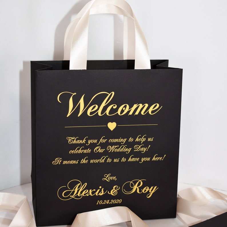 May include: Black gift bag with white satin handles. The bag has gold lettering that says "Welcome", "Thank you for coming to help us celebrate Our Wedding Day! It means the world to us to have you here! Love, Alexis & Roy 10.24.2020".