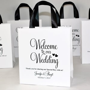 May include: White favour bags with black handles and the text "Welcome to our Wedding" with two small hearts. The bags also include the names "Jennifer & Raget" and the date "October 5, 2019".