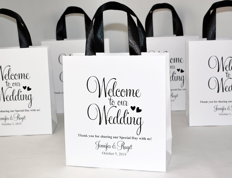 May include: White favour bags with black handles and the text "Welcome to our Wedding" with two small hearts. The bags also include the names "Jennifer & Raget" and the date "October 5, 2019".