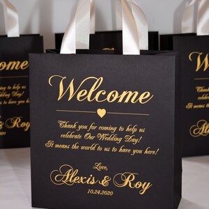 May include: Black gift bag with white satin handles and gold lettering. The bag says "Welcome" and "Thank you for coming to help us celebrate Our Wedding Day! It means the world to us to have you here! Love, Alexis & Roy 10.24.2020"