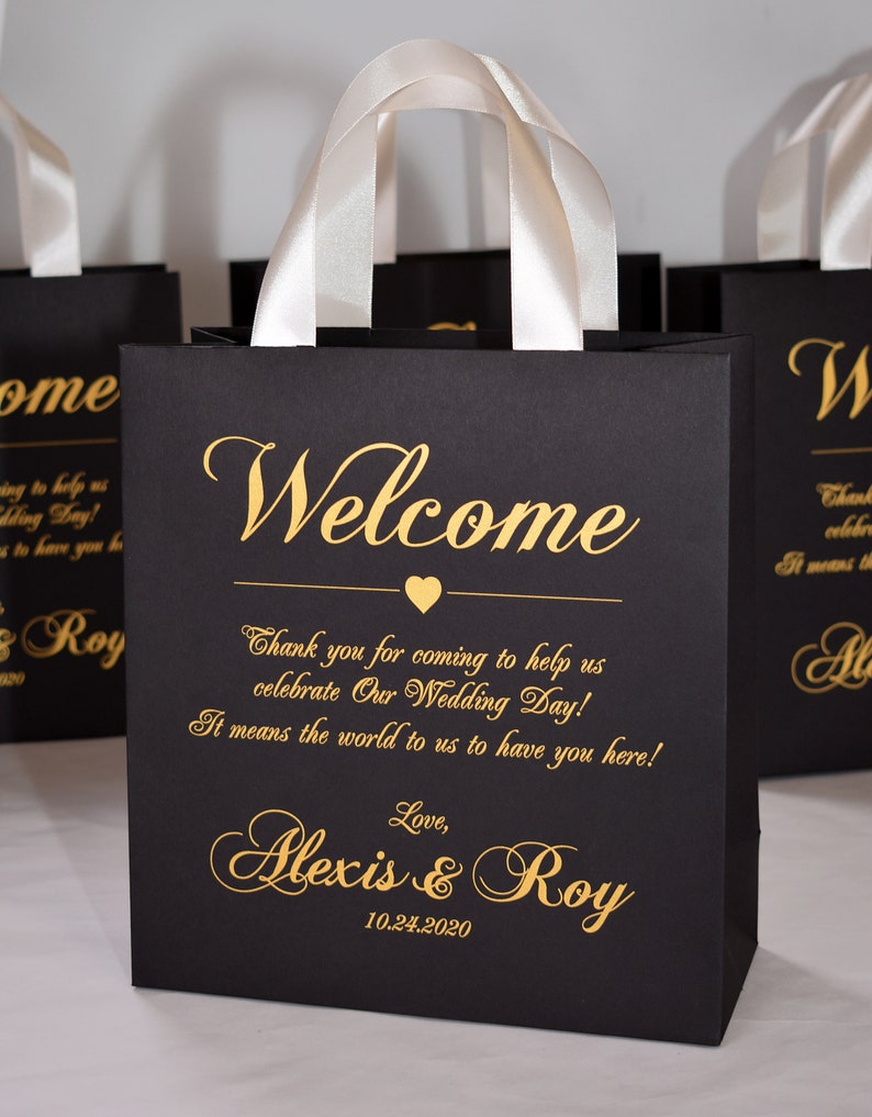 May include: Black gift bag with white satin handles and gold lettering. The bag says "Welcome" and "Thank you for coming to help us celebrate Our Wedding Day! It means the world to us to have you here! Love, Alexis & Roy 10.24.2020"