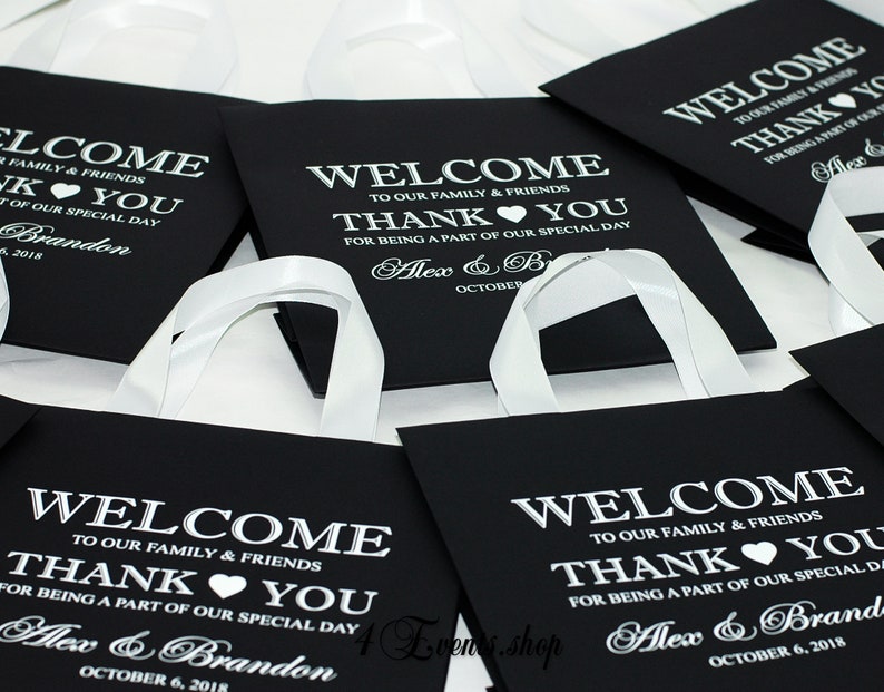 May include: Black favor bags with white satin ribbon handles. The bags have a welcome message, thank you message, and the names Alex & Brandon with the date October 6, 2018.