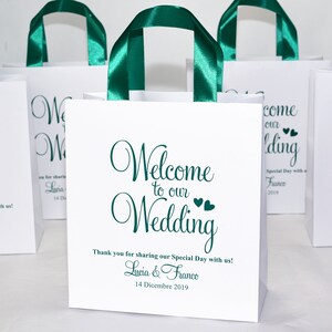 May include: White paper gift bags with green satin ribbon handles. The bags have a green script font that says "Welcome to our Wedding" with two small hearts. The text also includes "Thank you for sharing our Special Day with us! Lucia & Franco 14 Dicembre 2019."