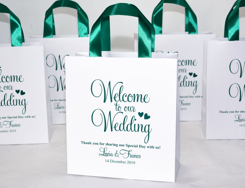 May include: White paper gift bags with green satin ribbon handles. The bags have a green script font that says "Welcome to our Wedding" with two small hearts. The text also includes "Thank you for sharing our Special Day with us! Lucia & Franco 14 Dicembre 2019."