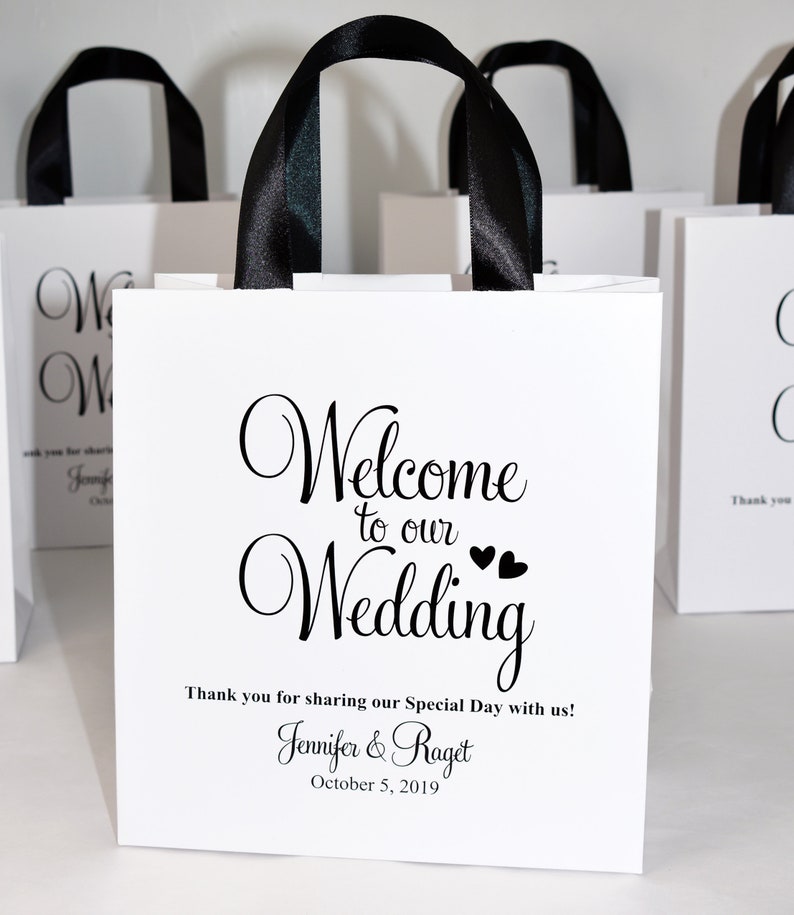 May include: White paper gift bag with black ribbon handles. The bag has a printed design that says "Welcome to our Wedding" with two small hearts. The text below says "Thank you for sharing our Special Day with us! Jennifer & Raget October 5, 2019"