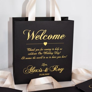 May include: Black and gold wedding favor bags with white satin handles. The bags have the text "Welcome" and "Thank you for coming to help us celebrate Our Wedding Day! It means the world to us to have you here! Love, Alexis & Roy 10.24.2020" printed on them.