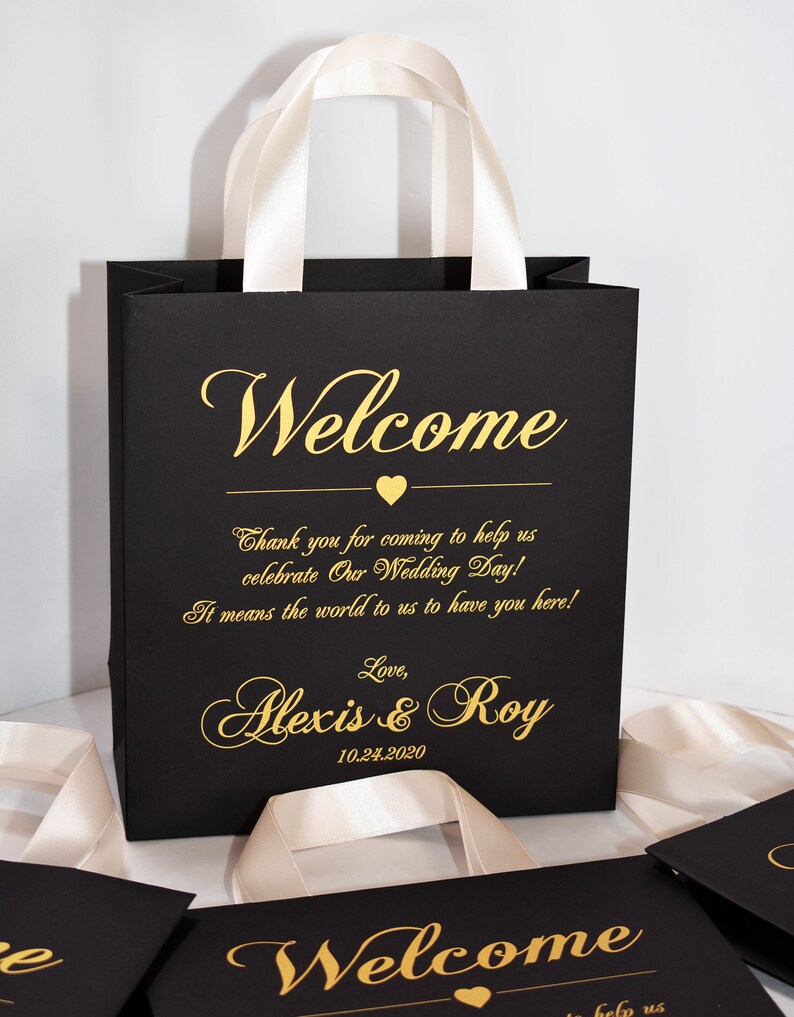 May include: Black and gold wedding favor bags with white satin handles. The bags have the text "Welcome" and "Thank you for coming to help us celebrate Our Wedding Day! It means the world to us to have you here! Love, Alexis & Roy 10.24.2020" printed on them.