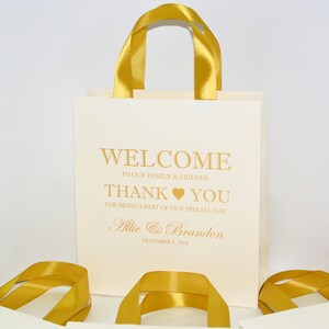 May include: White favor bags with gold ribbon handles. The bags have a gold inscription that reads "Welcome to our family & friends Thank you for being a part of our special day Allie & Brandon December 8, 2013".