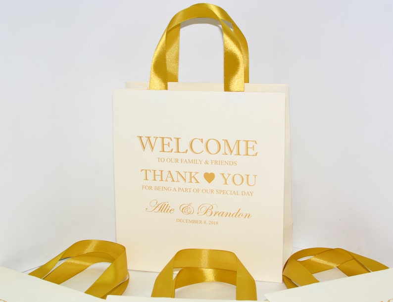 May include: White favor bags with gold ribbon handles. The bags have a gold inscription that reads "Welcome to our family & friends Thank you for being a part of our special day Allie & Brandon December 8, 2013".