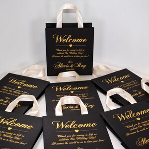 May include: Black gift bags with white satin handles and gold lettering. The bags say "Welcome" and "Thank you for coming to help us celebrate Our Wedding Day! It means the world to us to have you here! Love Alexis & Roy 10.24.2020".