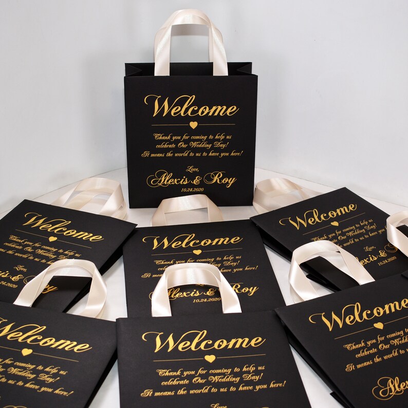 May include: Black gift bags with white satin handles and gold lettering. The bags say "Welcome" and "Thank you for coming to help us celebrate Our Wedding Day! It means the world to us to have you here! Love Alexis & Roy 10.24.2020".