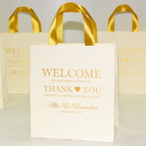 May include: White favor bags with gold handles and gold lettering that reads "Welcome to our family & friends Thank you for being a part of our special day Allie & Brandon December 8, 2018".