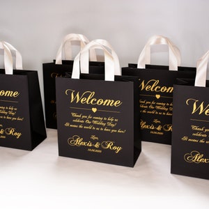 May include: Black gift bags with white satin handles and gold lettering. The bags say "Welcome" and "Thank you for coming to help us celebrate Our Wedding Day! It means the world to us to have you here! Love, Alexis & Roy 10.24.2020".