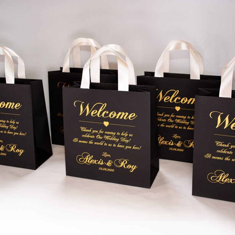 May include: Black gift bags with white satin handles and gold lettering. The bags say "Welcome" and "Thank you for coming to help us celebrate Our Wedding Day! It means the world to us to have you here! Love, Alexis & Roy 10.24.2020".