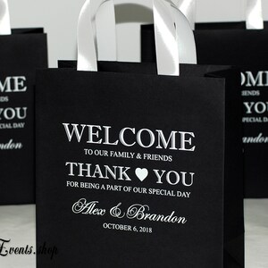 May include: Black paper gift bags with white satin handles and white text. The text reads "Come Emily & Friends, K You, Part of our special day, Alex & Brandon, October 6, 2018, Welcome to our family & friends, Thank You for being a part of our special day, Alex & Brandon, October 6, 2018".