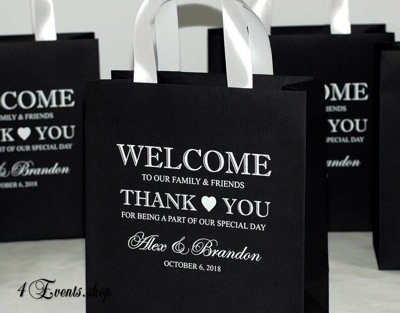 May include: Black paper gift bags with white satin handles and white text. The text reads "Come Emily & Friends, K You, Part of our special day, Alex & Brandon, October 6, 2018, Welcome to our family & friends, Thank You for being a part of our special day, Alex & Brandon, October 6, 2018".