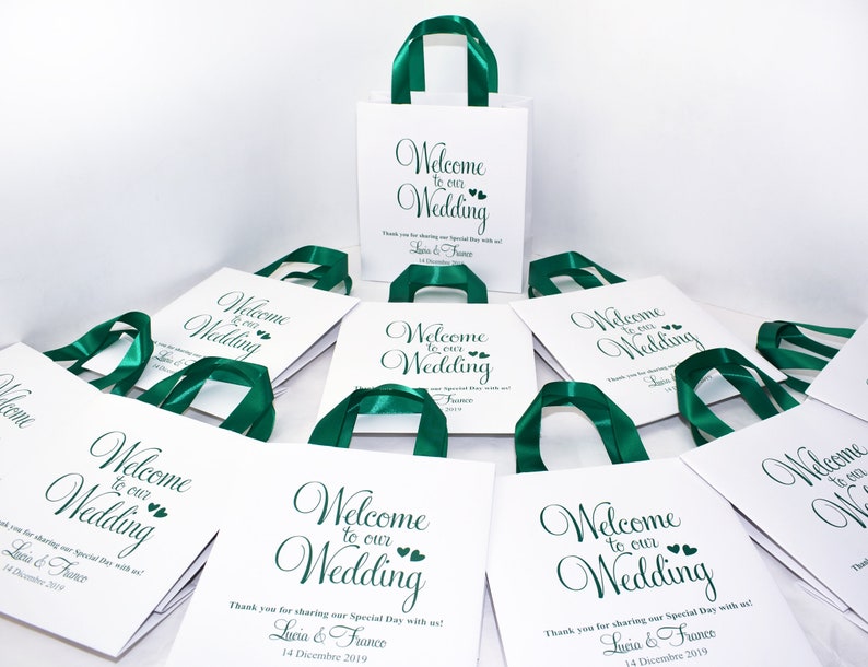 May include: White gift bags with green satin ribbon handles. The bags have a printed message that says "Welcome to our Wedding" with the names Lucia & France and the date 14 December 2019.