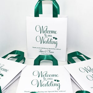 May include: White gift bags with green satin ribbon handles. The bags have the text "Welcome to our Wedding" printed on them. The bags are decorated with small hearts. The text "Thank you for sharing our Special Day with us!" is printed below the main text. The bags are personalized with the names "Lucia & Franco" and the date "14 Dicembre 2019".