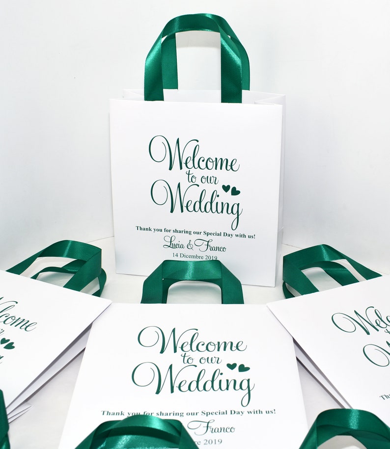 May include: White gift bags with green satin ribbon handles. The bags have the text "Welcome to our Wedding" printed on them. The bags are decorated with small hearts. The text "Thank you for sharing our Special Day with us!" is printed below the main text. The bags are personalized with the names "Lucia & Franco" and the date "14 Dicembre 2019".