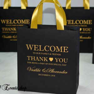 May include: Black gift bags with gold ribbon handles and gold lettering. The bags say "Welcome to our family & friends, Thank you for being a part of our special day, Vasiliki & Alexander, December 8, 2018".