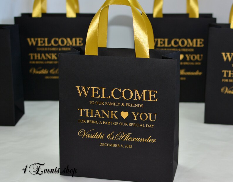 May include: Black gift bags with gold ribbon handles and gold lettering. The bags say "Welcome to our family & friends, Thank you for being a part of our special day, Vasiliki & Alexander, December 8, 2018".
