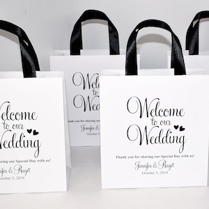 May include: White gift bags with black ribbon handles and the text "Welcome to our Wedding" printed on the front. The bags are decorated with small hearts and include the names Jennifer & Raget and the date 5 October 2019.