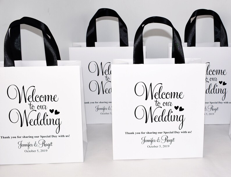 May include: White gift bags with black ribbon handles and the text "Welcome to our Wedding" printed on the front. The bags are decorated with small hearts and include the names Jennifer & Raget and the date 5 October 2019.