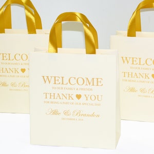 May include: Three white gift bags with gold handles and gold lettering. The bags say "Welcome to our family & friends, Thank You for being a part of our special day, Allie & Brandon, December 8, 2018".