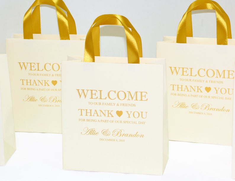 May include: Three white gift bags with gold handles and gold lettering. The bags say "Welcome to our family & friends, Thank You for being a part of our special day, Allie & Brandon, December 8, 2018".