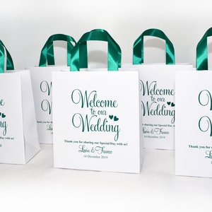 May include: White paper gift bags with teal ribbon handles. The bags have the text "Welcome to our Wedding" printed on them with a small heart design. The text also includes the names "Lucia & France" and the date "14 Dicembre 2019".