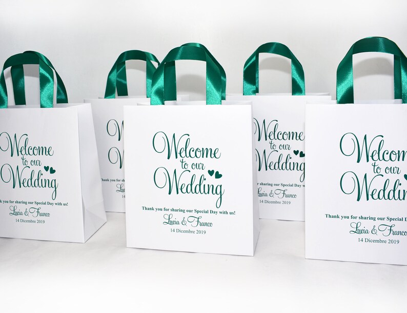 May include: White paper gift bags with teal ribbon handles. The bags have the text "Welcome to our Wedding" printed on them with a small heart design. The text also includes the names "Lucia & France" and the date "14 Dicembre 2019".