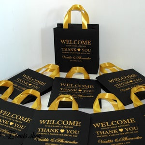 May include: Black and gold favor bags with gold ribbon handles. The bags have a welcome message and thank you message with the names Vasiliki & Alexander and the date December 8, 2018.