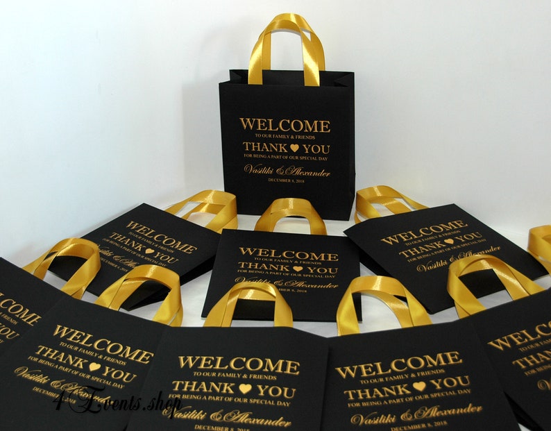 May include: Black and gold favor bags with gold ribbon handles. The bags have a welcome message and thank you message with the names Vasiliki & Alexander and the date December 8, 2018.