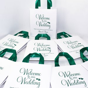 May include: White gift bags with green satin ribbon handles. The bags have a green script font that says "Welcome to our Wedding" with two small hearts. The bags have a thank you message and the couple's names and wedding date.