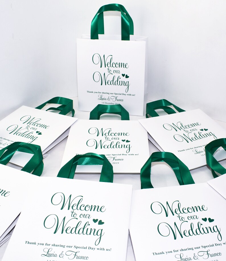 May include: White gift bags with green satin ribbon handles. The bags have a green script font that says "Welcome to our Wedding" with two small hearts. The bags have a thank you message and the couple's names and wedding date.