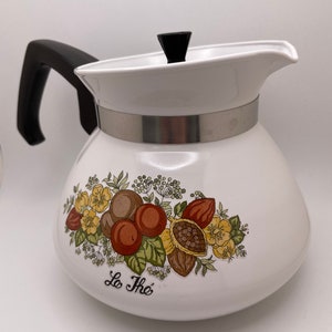 May include: White teapot with a black handle and lid. The teapot has a silver band around the middle and a floral design with the words "Le The" on the front.