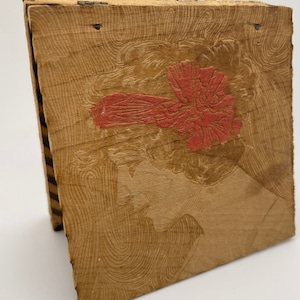 Può includere: Una scatola di legno con un coperchio incernierato. La scatola è decorata con un'immagine rossa e bianca di un viso di donna.