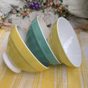 Peut inclure: Trois bols en céramique jaune, vert d'eau et blanc, disposés sur une surface à carreaux jaunes et blancs. Les bols ont un extérieur nervuré et un intérieur lisse et blanc. Les bols sont empilés en biais.