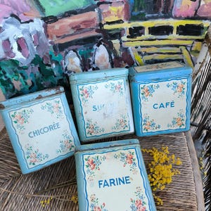 Può includere: Quattro barattoli vintage in metallo blu con disegni floreali ed etichette con la scritta "CHICOREE", "SUCRE", "CAFÉ" e "FARINE". I barattoli hanno una forma rettangolare e sono disposti su una superficie tessuta.