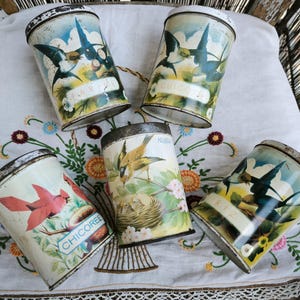 Peut inclure: Cinq boîtes métalliques vintage avec des illustrations d'oiseaux. Les boîtes présentent des images d'hirondelles, d'un cardinal et d'un oiseau avec un nid. Chaque boîte porte un nom différent, comme "Chicoree" et "Farine". Les boîtes sont sur un tissu blanc brodé.