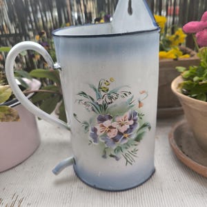 May include: Vintage white enamel wall planter with a blue rim and handle. The planter features a floral design with purple, pink, and yellow flowers. It has a spout at the bottom and a hole for hanging.