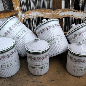 Può includere: Un set di barattoli da cucina in ceramica bianca con coperchi. Ogni barattolo presenta un motivo floreale e un'etichetta verde con testo francese. I barattoli sono disposti su una superficie di legno, evocando uno stile vintage o rustico.