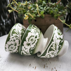 Op de afbeelding: Drie gestapelde keramische kommen met een groen en wit bloemmotief. De kommen staan op een wit houten oppervlak, met een plant in een pot op de achtergrond. De kommen hebben een voet en een gebogen rand.