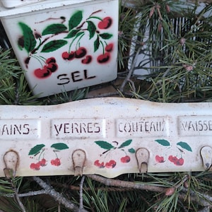 Può includere: Un organizer da cucina vintage in metallo bianco con ganci, con le parole "MAINS, VERRES, COUTEAUX, VAISSELLE". È decorato con ciliegie rosse e foglie verdi. È visibile anche un contenitore abbinato con "SEL".