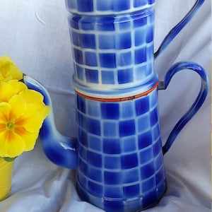 Può includere: Una caffettiera vintage in smalto blu e bianco con un motivo a scacchi. La caffettiera ha un coperchio, un beccuccio e un manico. La caffettiera è decorata con un bordo rosso. Fiori gialli in un vaso giallo sullo sfondo.